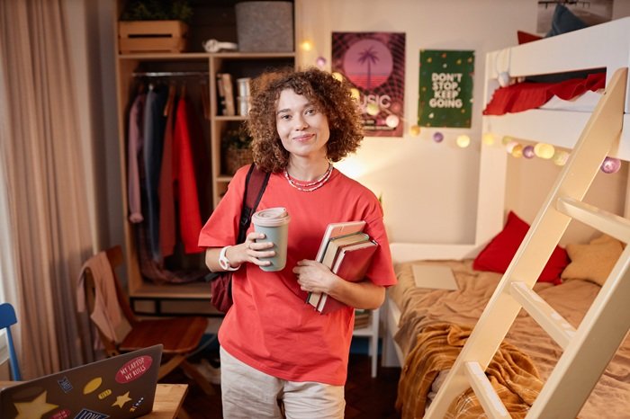 Waist up portrait of curly haired girl as female student smiling at camera and holding books in college dorm, copy space